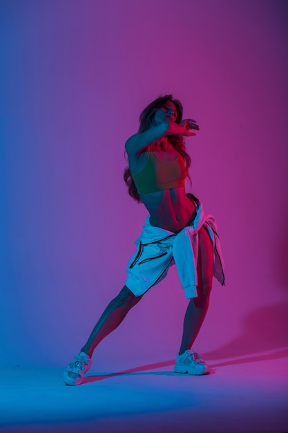 Young woman dancer in stylish youth clothes in gym shoes dancing and posing in a room with bright neon blue-pink color. Sports girl enjoys a dance in the studio with multi-colored ultraviolet light.