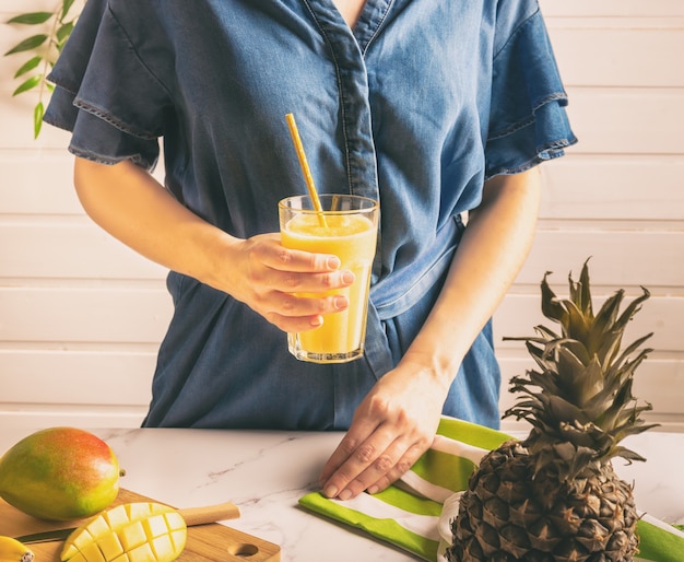 Young woman in denim dress holding a glass with mango and pineapple smoothie in her hand. Healthy lifestyle.