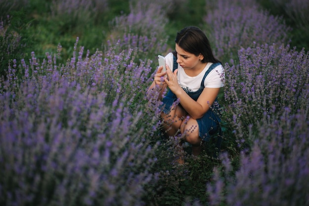 Young woman in denim dress sitting in lavender field in summer Beautiful girl smile and make photo