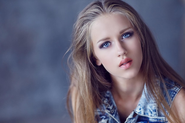 A young woman in denim jacket and black pants posing near concrete wall.