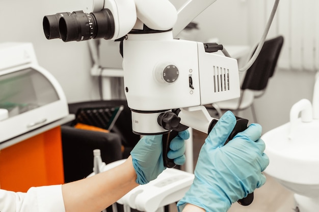 Young woman dentist doctor looks through a professional microscope in a dental clinic. A doctor in a disposable medical mask and cap. Advanced equipment in dentistry