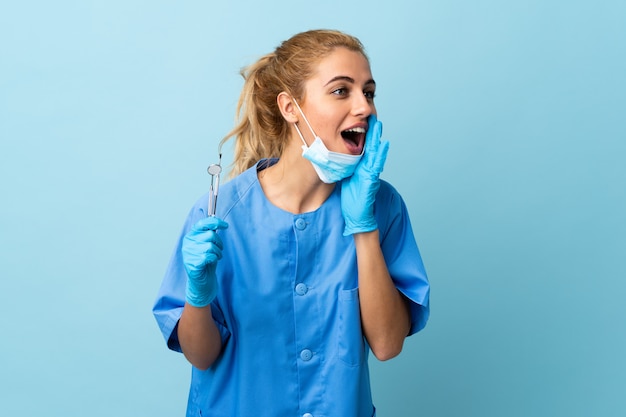 Young woman dentist holding tools over blue wall shouting with mouth wide open