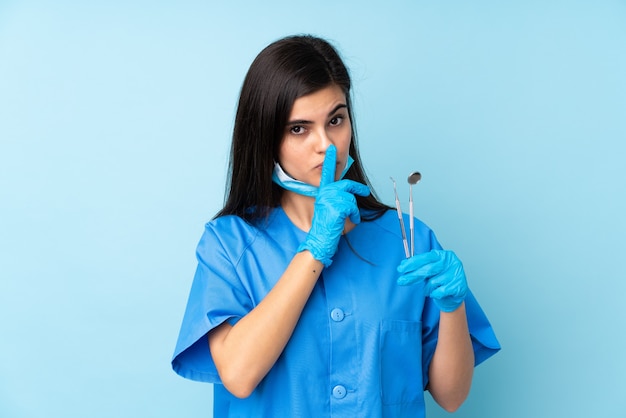 沈黙のジェスチャーをしている孤立した青い壁の上にツールを保持している若い女性の歯科医