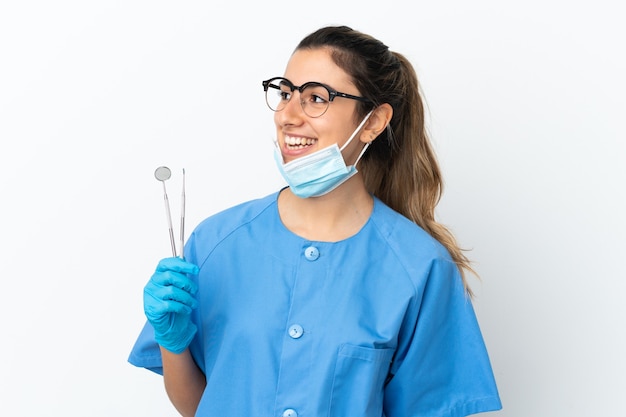 Young woman dentist holding tools isolated on white background looking to the side and smiling