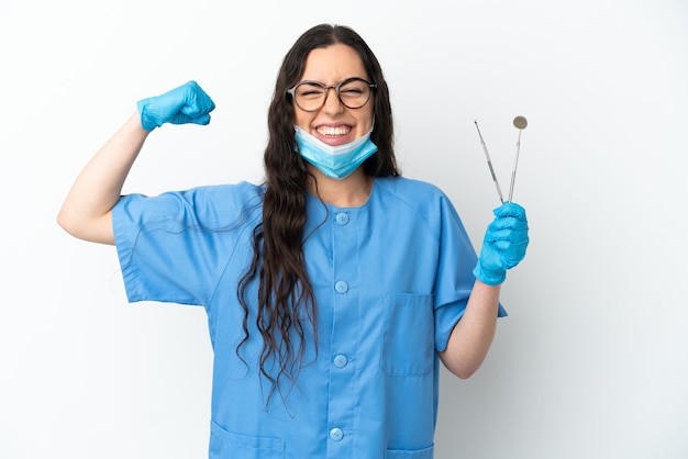 Young woman dentist holding tools isolated on white wall doing strong gesture