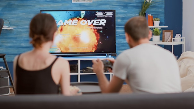 Young woman disappointed because of gamer boyfriend losing online game while playing on console using wireless controller. Couple arguing over failed level in space shooter in front of big screen tv.