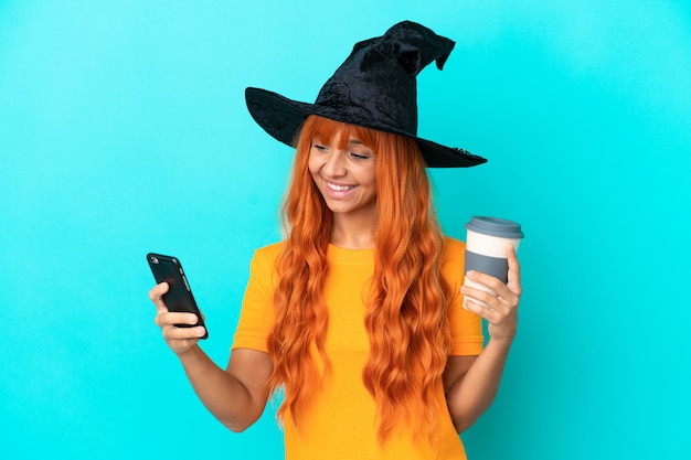 Young woman disguised as witch isolated on blue background holding coffee to take away and a mobile