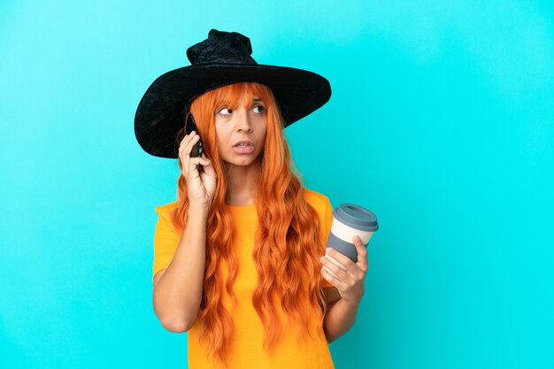 Young woman disguised as witch isolated on blue background holding coffee to take away and a mobile