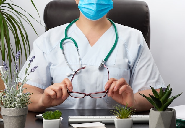 Young woman doctor in a blue uniform, a disposable medical mask sits in a chair at the table and works