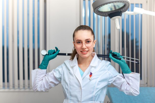 Young woman doctor in a coat with a phonendoscope in hands posing at the camera on the background of the ward in the hospital. Copy Space, Medecine Concept.