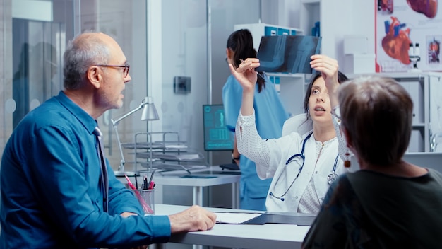 Young woman doctor getting X Ray from nurse while talking with old couple about their problems. Modern hospital or private clinic healthcare checkup for disease prevention and healthcare problems. Pat