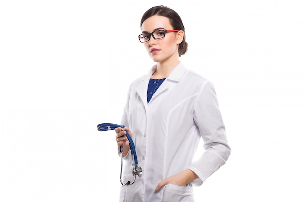 Young woman doctor holding stethoscope in one hand and her other hand in pocket in white uniform on white background