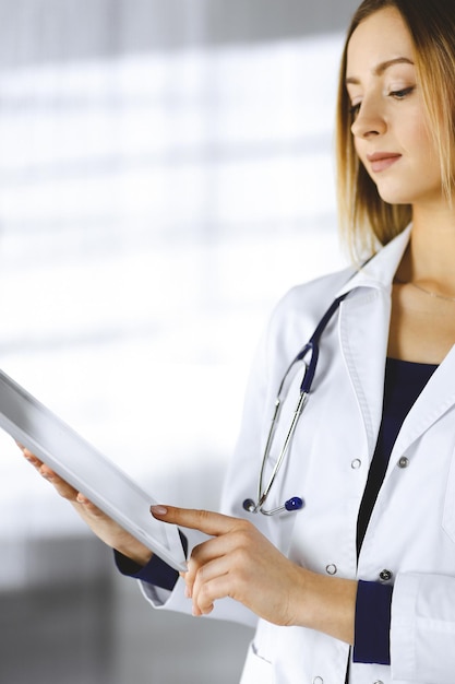 Young woman-doctor is holding a tablet computer in her hands, while standing in a clinic. Portrait of friendly female physician with a stethoscope at work. Perfect medical service in a hospital. Medic