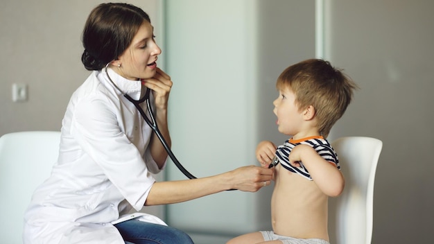 Young woman doctor listening little boy with stethoscope in medical office