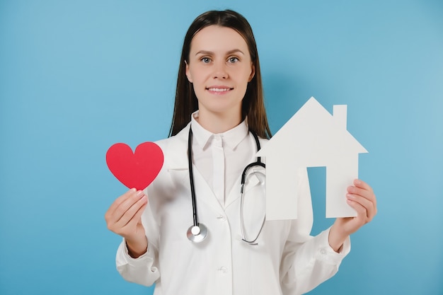 Il medico della giovane donna in uniforme bianca medica professionale e lo stetoscopio tiene la piccola casa e il cuore rosso, felice che esamina macchina fotografica, posante sopra il fondo blu dello studio. cura, medicina, aiuto e supporto