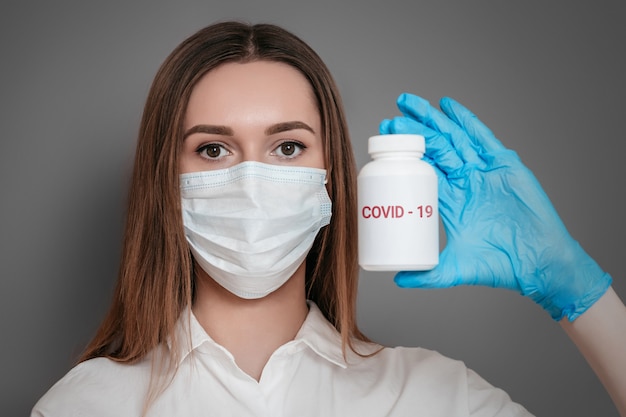 A young woman doctor virologist in a protective medical mask on her face, holding a jar of pills with covid-19 words isolated over dark grey background. Coronavirus concept