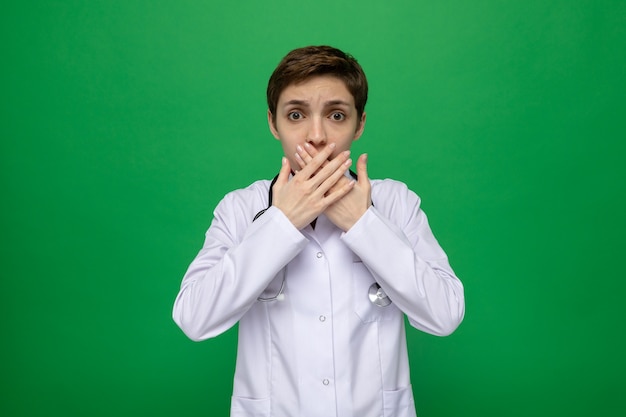 Молодая женщина-врач в белом халате со стетоскопом смотрит в шоке, прикрывая рот руками