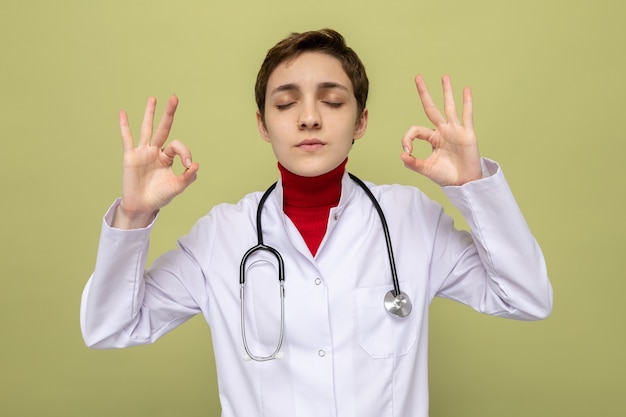 Молодая женщина-врач в белом халате со стетоскопом делает жест медитации, пытаясь расслабиться с закрытыми глазами