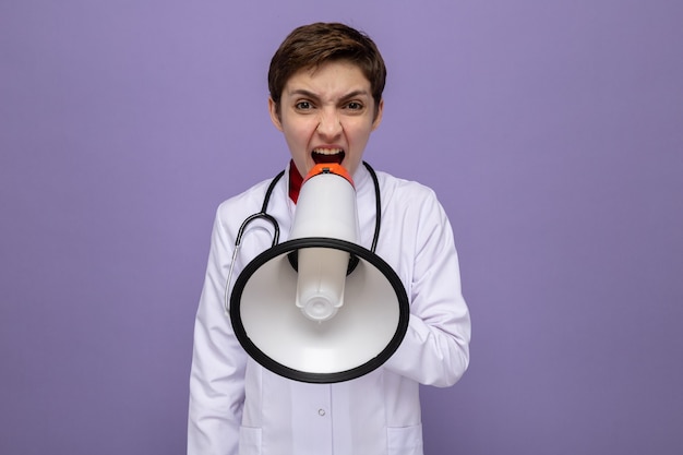 Young woman doctor in white coat with stethoscope shouting to megaphone angry and excited