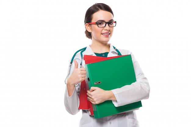 Young woman doctor with stethoscope holding binders in her hands with thumb up in white uniform on white 