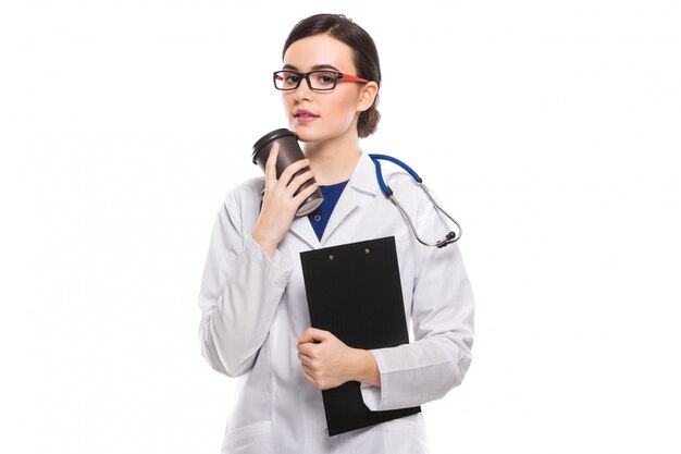 Young woman doctor with stethoscope holding clipboard and cup of coffee in her hands in white uniform