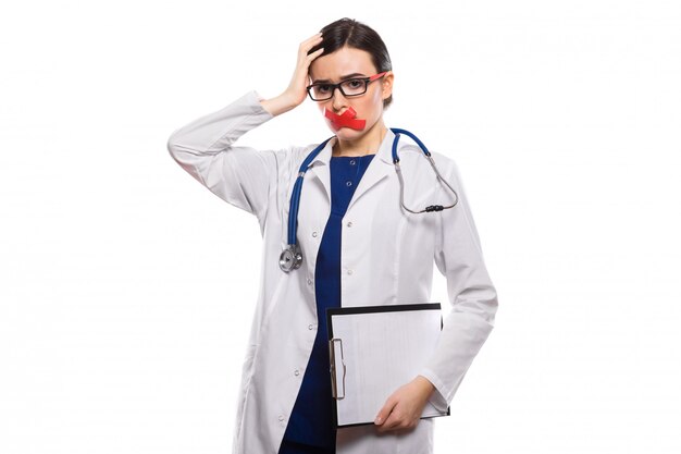 Young woman doctor with stethoscope holding clipboard in her hands with taped lips in white uniform on white 