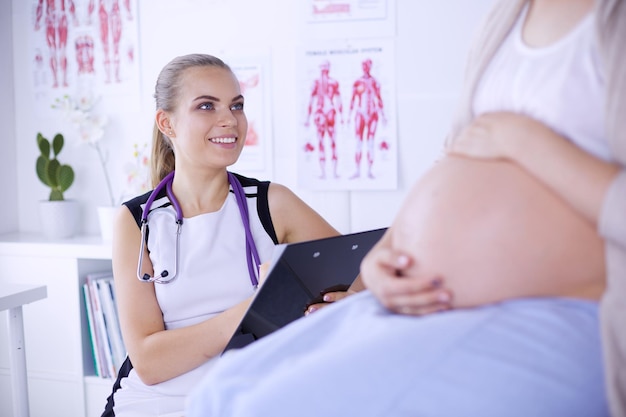 Young woman doctor with stethoscope and tablet speaking with pregnant woman at hospital