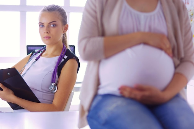 Young woman doctor with stethoscope and tablet speaking with pregnant woman at hospital