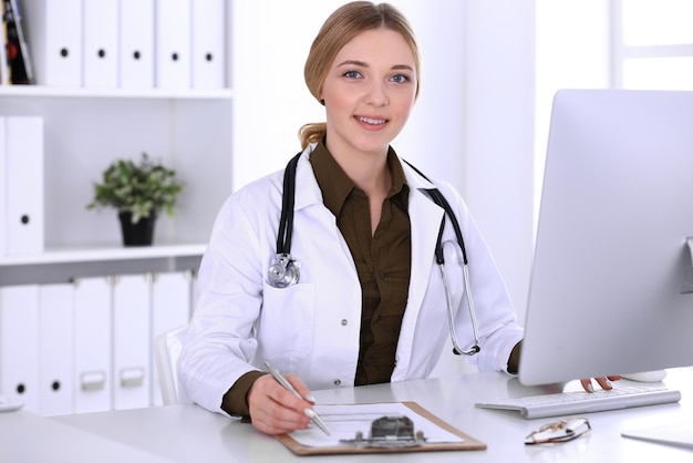 Young woman doctor at work in hospital looking at desktop pc monitor. Physician controls medication history records and exam results. Medicine and healthcare concept.