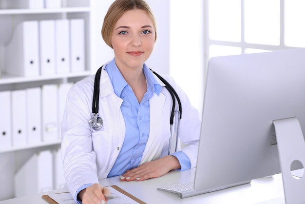Young woman doctor at work in hospital looking at desktop pc monitor. Physician controls medication history records and exam results. Medicine and healthcare concept.