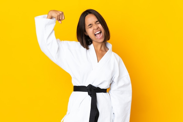 Young woman doing karate isolated on yellow background doing strong gesture