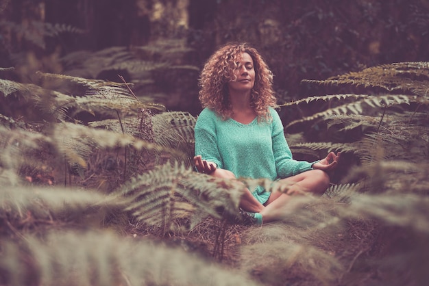 Young woman doing meditation by sitting in forest with eyes closed. Caucasian woman doing yoga for fitness at park. Carefree woman meditating in lotus position amidst plants in forest