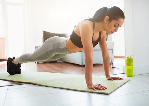 Young woman doing sport workout in room during quarantine. Concentrated calm girl stand in plank position using hands. Look down with concentration.