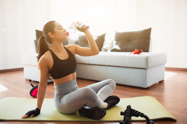 Young woman doing sport workout in room during quarantine. Rest after exercise. Girl sit on mat and drink water from plastic bottle. Pause after wokrout.