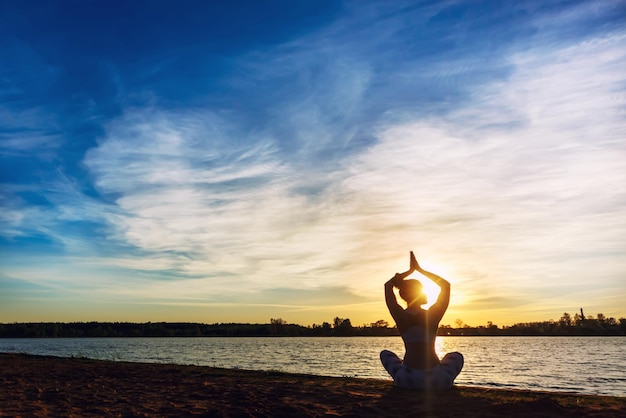 Young woman doing yoga exercises on the lake beach at sunset