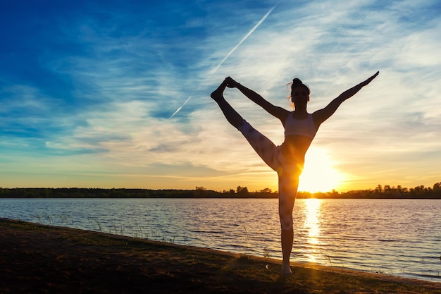 Young woman doing yoga exercises on the lake beach at sunset