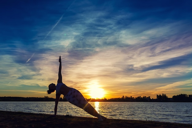 Young woman doing yoga exercises on the lake beach at sunset