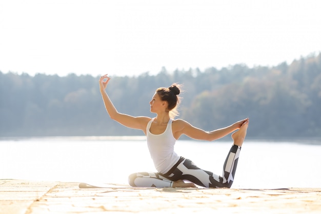 Young woman doing yoga near lake outdoors, meditation. Sport fitness and exercising in nature. Autumn sunset.