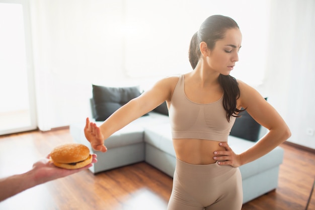 Young woman doing yoga workout in room during quarantine. Girl refuses to eat burger.