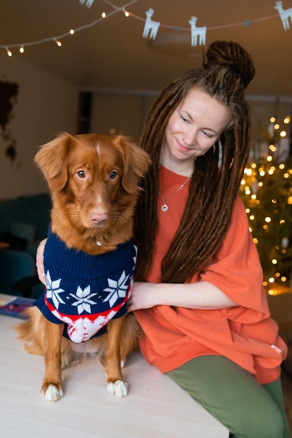 Фото Молодая женщина с дредами проводит время со своей собакой в свитере дома на праздниках