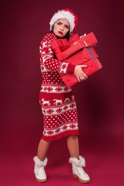 Young woman in dress and christmas hat holding gift boxes and looking at the camera over red background