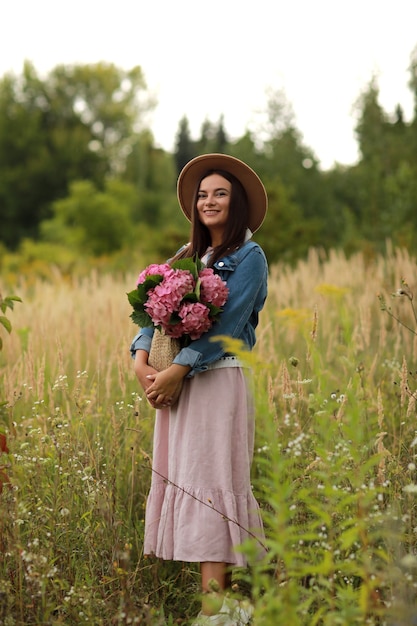 Young woman in dress, hat and denim jacket holding bouquet of pink flowers hydrangea,