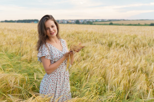 Young woman in dress holds wheat ears in hands, summer field. Smiling girl on golden meadow. Freedom