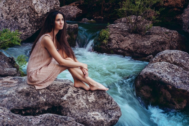 Young woman in the dress is sitting on the stone in the middle of a creek Fashionable toning