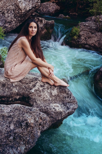 Young woman in the dress is sitting on the stone in the middle of a creek Fashionable toning