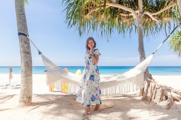 Young woman in dress standing near hammock between palm trees