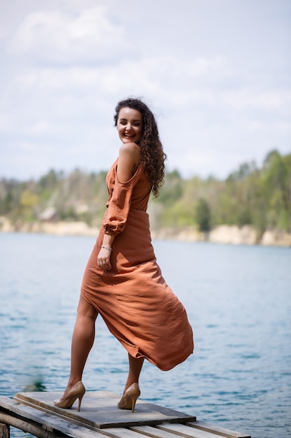 Una giovane donna in abito si erge su una muratura di legno nel mezzo di un lago blu. sorrisi felici della ragazza e il sole splende, giorno d'estate. ha i capelli lunghi ricci e un aspetto europeo