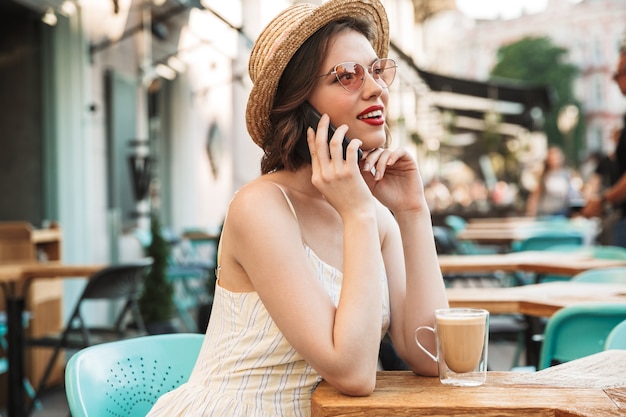 Giovane donna in abito e cappello di paglia parlando di smartphone