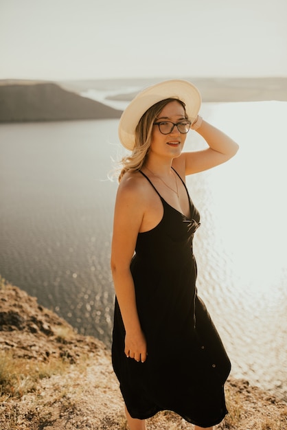 帽子と眼鏡をかけた若い女性のドレスは、日没時に海の上の崖の上に立っています。