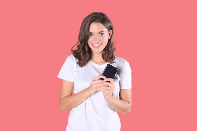 Young woman dressed in basic clothing isolated on pink background using mobile phone